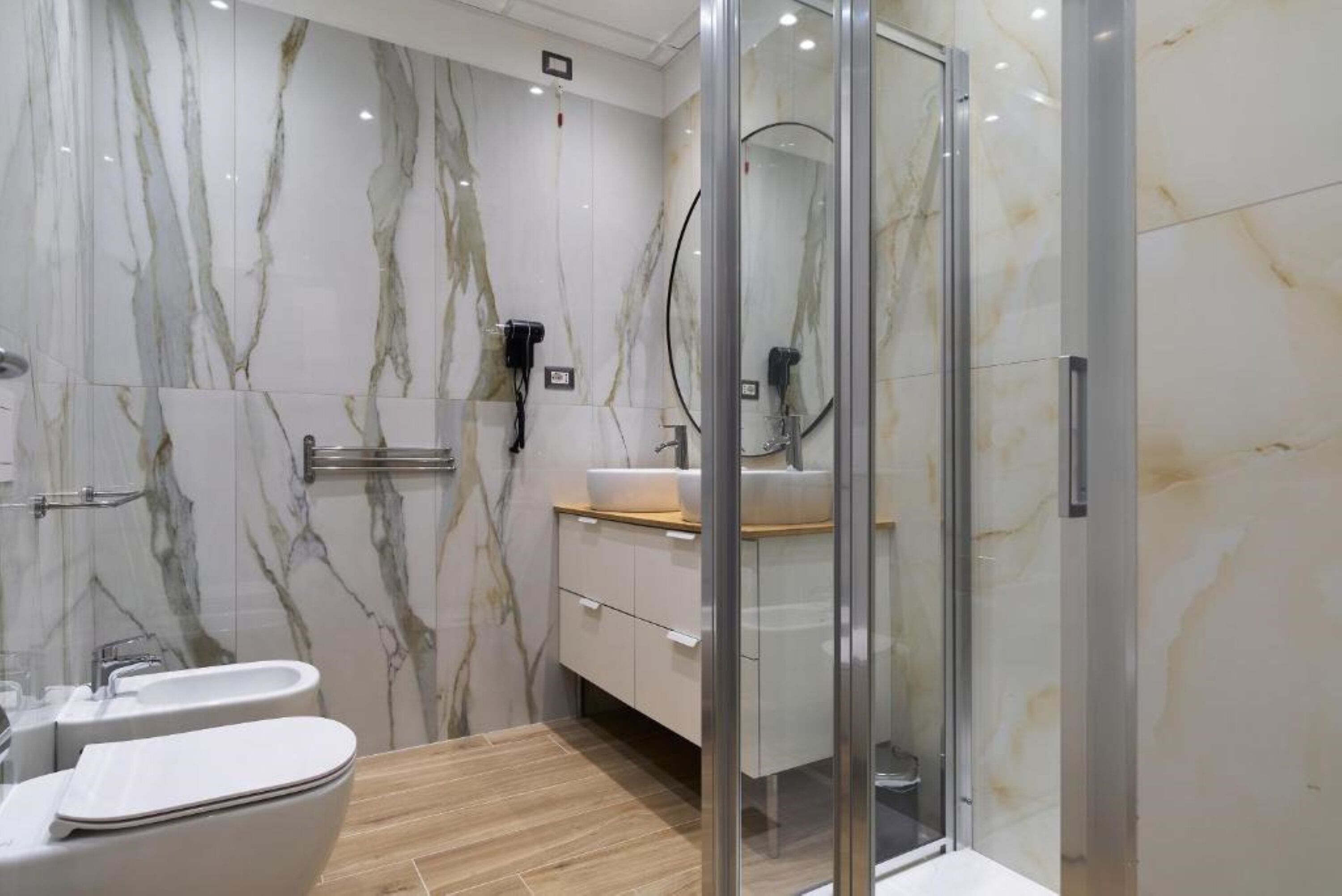 Une salle de bain moderne avec des murs en marbre, une douche en verre, un lavabo blanc et un plancher en bois.