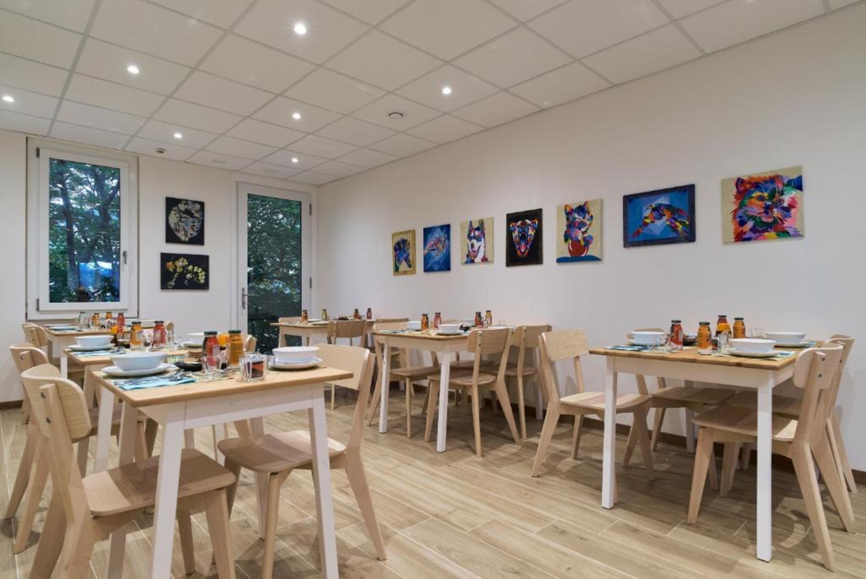 A dining area with multiple tables and chairs, artwork on the walls, and large windows.