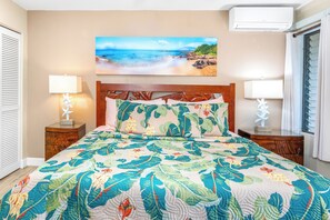 Une chambre à coucher comprenant un lit, deux tables de chevet, un climatiseur fixé au mur et un tableau sur le thème de la plage au-dessus du lit.