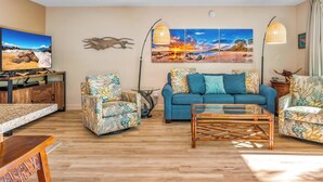 Ein Wohnzimmer mit einem blauen Sofa, floralen Ohrensesseln, einem Glastisch, einem Fernseher auf einem Holzboden und Kunstwerken an der Wand.