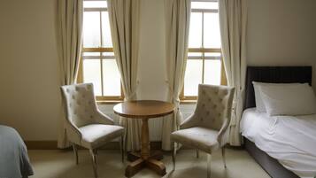 Une chambre d’hôtel avec un lit, deux fauteuils, une table ronde en bois et une fenêtre avec des rideaux.