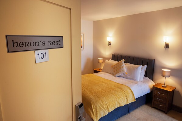 Una habitación de hotel con una cama, dos mesitas de noche con lámparas, un cartel en la pared que dice "heron's rest 101" y una puerta.