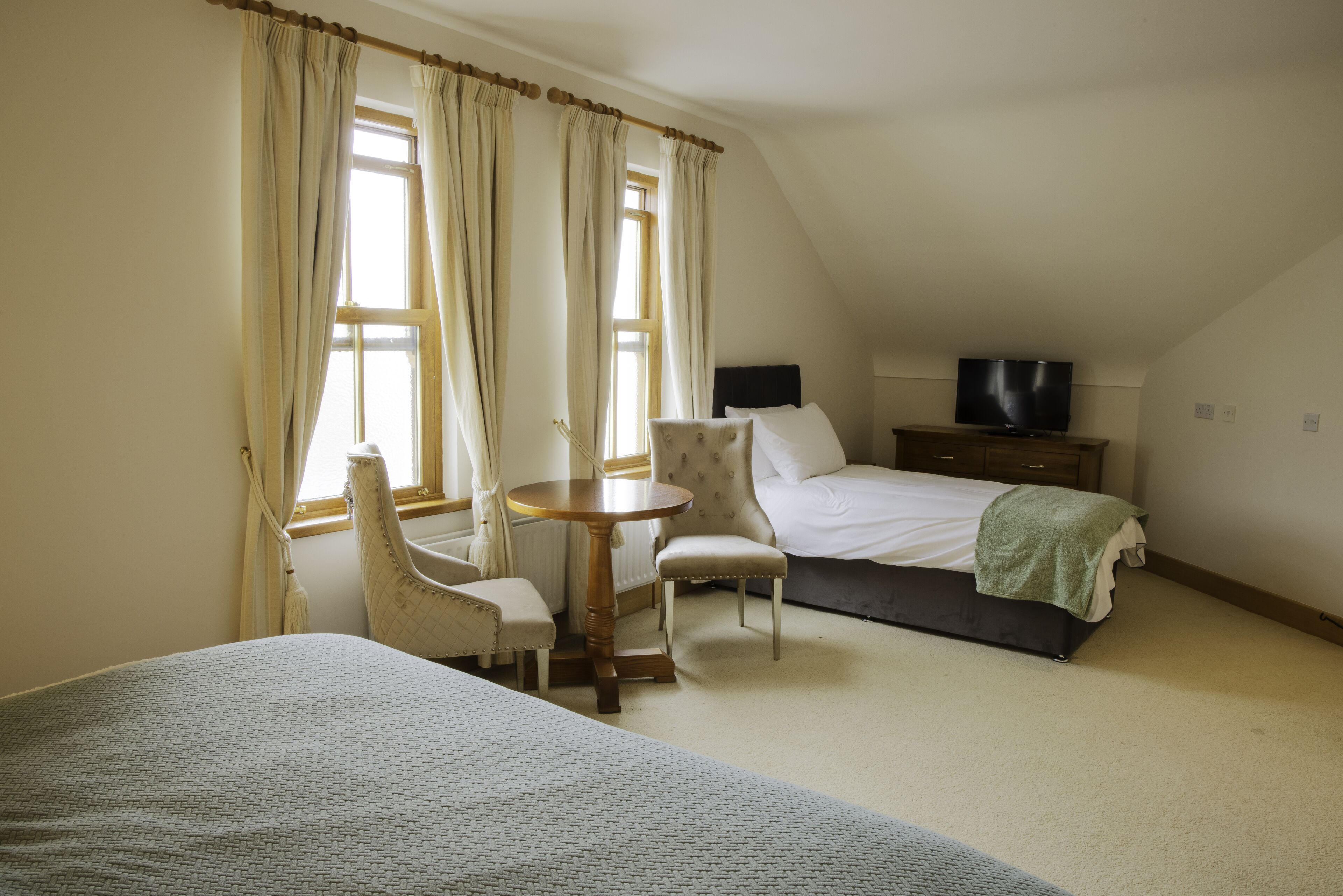 Habitación de hotel con cama, televisor, una mesita redonda, dos sillas y ventana con cortinas.