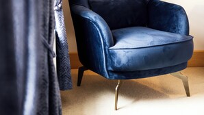 Een donkerblauwe fluweel fauteuil met metalen poten, geplaatst in een kamer met een lichtkleurig tapijt en een ingelijst kunstwerk aan de muur.