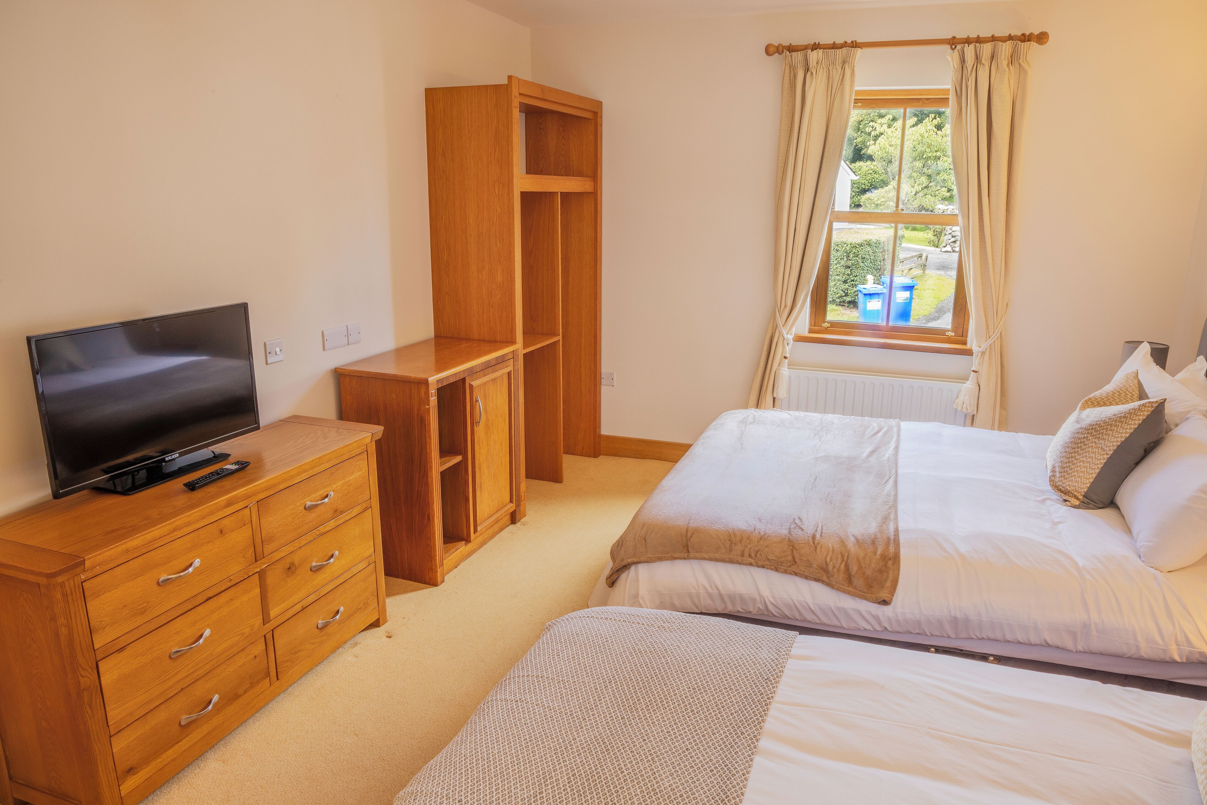 A bedroom with a TV on a wooden dresser, a wardrobe, a bed with pillows, and a window with curtains.