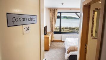 A hotel room with a bed, a dresser, a television, and a window with a view of a body of water and hills.