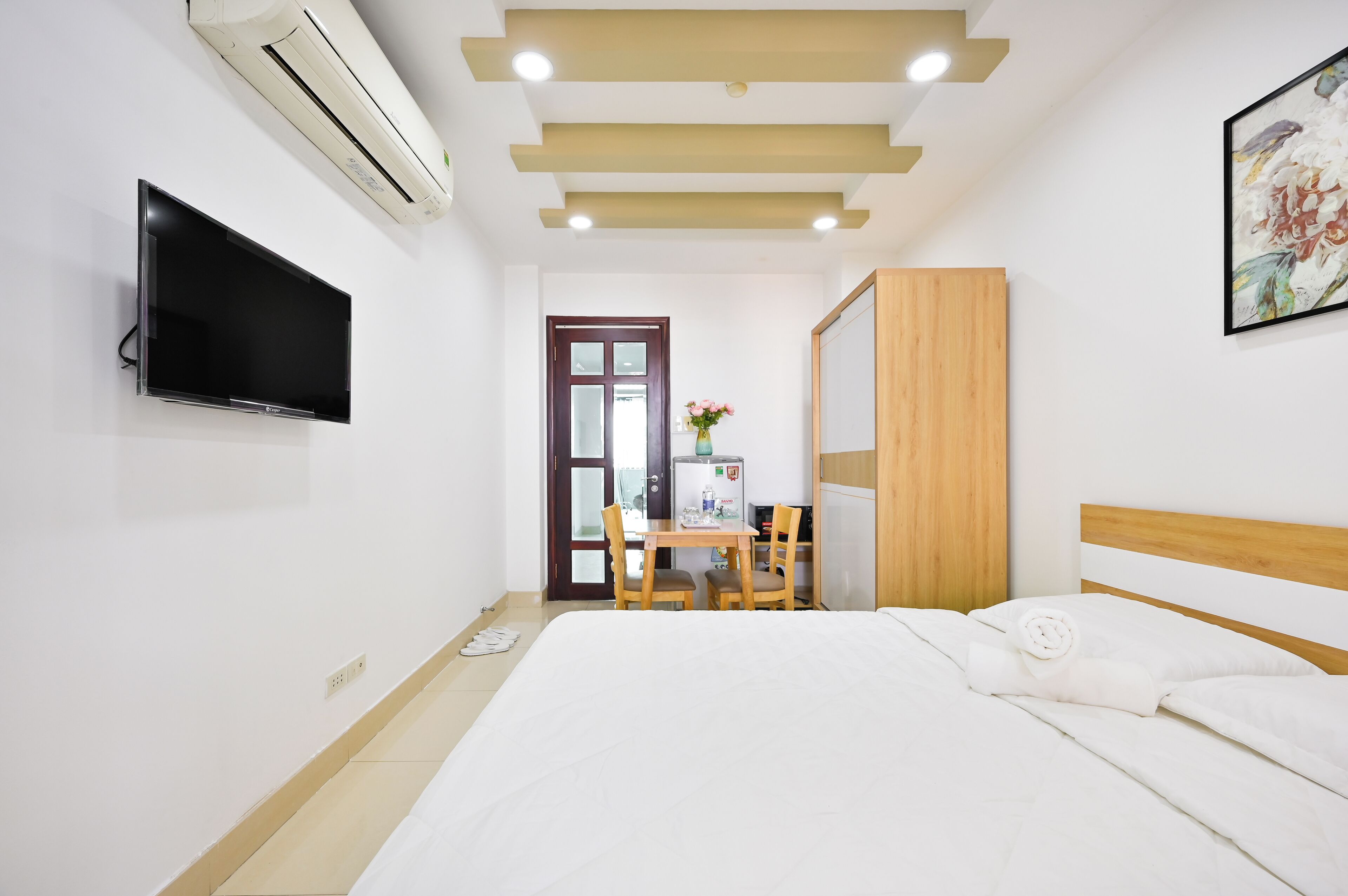 A hotel room with a bed, a desk with a chair, a TV mounted on the wall, and a door leading to the outside.