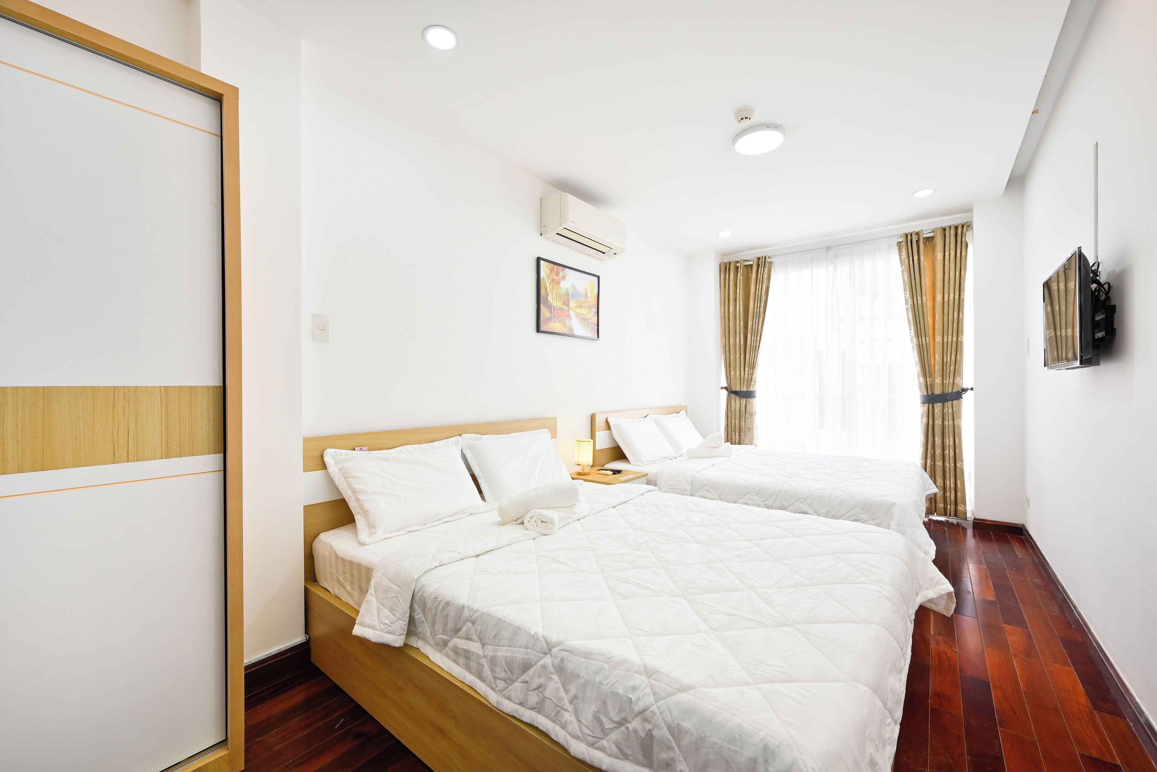 A hotel room with two beds, a wooden wardrobe, a wall-mounted air conditioner, a bedside table with a lamp, and a window with curtains.