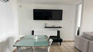 Una sala de estar moderna con una mesa de comedor de vidrio, sillas blancas, un sofá negro y un televisor de pantalla plana montado en la pared.