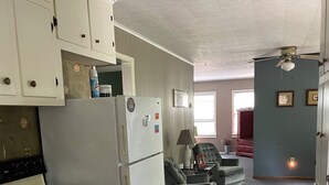 Cocina compacta con estufa, refrigerador y armarios. Hay una zona de comedor con un sofá y una lámpara. El piso está alfombrado y tiene un ventilador de techo.