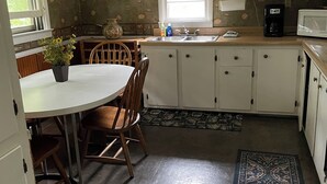 Una cocina con una mesa de comedor, sillas, un reloj, una ventana con persianas y un microondas.