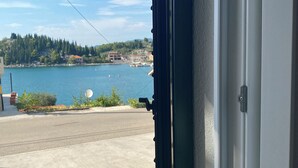 Blick durch ein Fenster mit Metallrahmen, eine Betonstraße und ein Gewässer mit Gebäuden und Grün im Hintergrund.