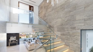 Un intérieur moderne avec un escalier en verre, une vue sur la ville depuis l’étage supérieur et un coin salon avec des canapés et une table basse.