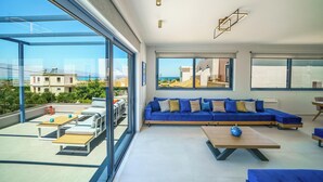Una sala de estar moderna con un sofá azul, una mesa de centro de madera y ventanas amplias que ofrecen vistas al mar y edificios.