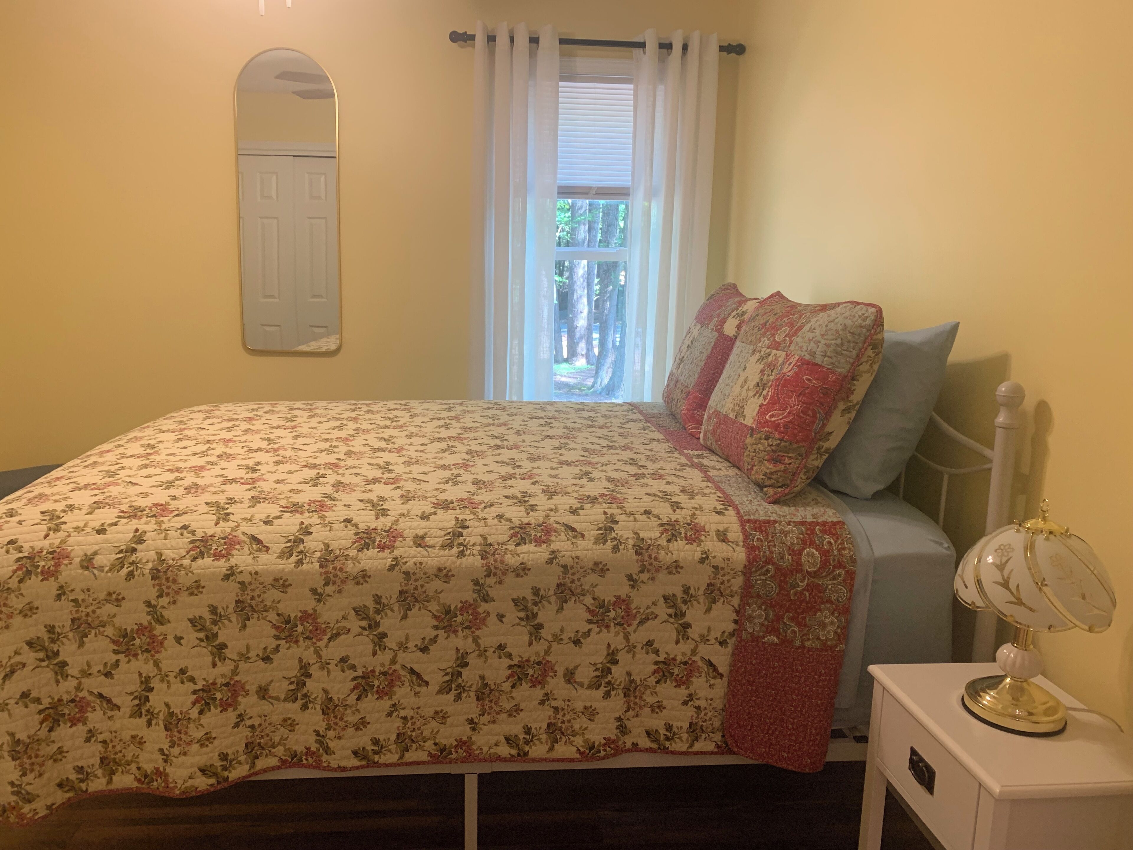 A bedroom with a floral bedspread, a window with curtains, a bedside table with a lamp, and a mirror on the wall.