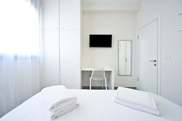 A hotel room with a bed, a TV mounted on the wall, a desk with a chair, and a wardrobe.