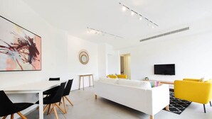 Una sala de estar moderna con un sofá blanco, sillones amarillos, una mesa de comedor con sillas negras y un cuadro abstracto grande en la pared.
