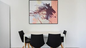 Un comedor con una mesa blanca y sillas negras, un cuadro abstracto enmarcado en la pared, y una pared blanca lisa.
