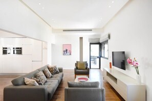 A modern living room with a grey sofa, a small table, and a TV on a white console. A kitchen with white cabinets and appliances is visible in the background.