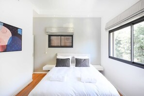 Un dormitorio moderno con una cama grande, una ventana con persianas, un equipo de aire acondicionado y un cuadro en la pared.