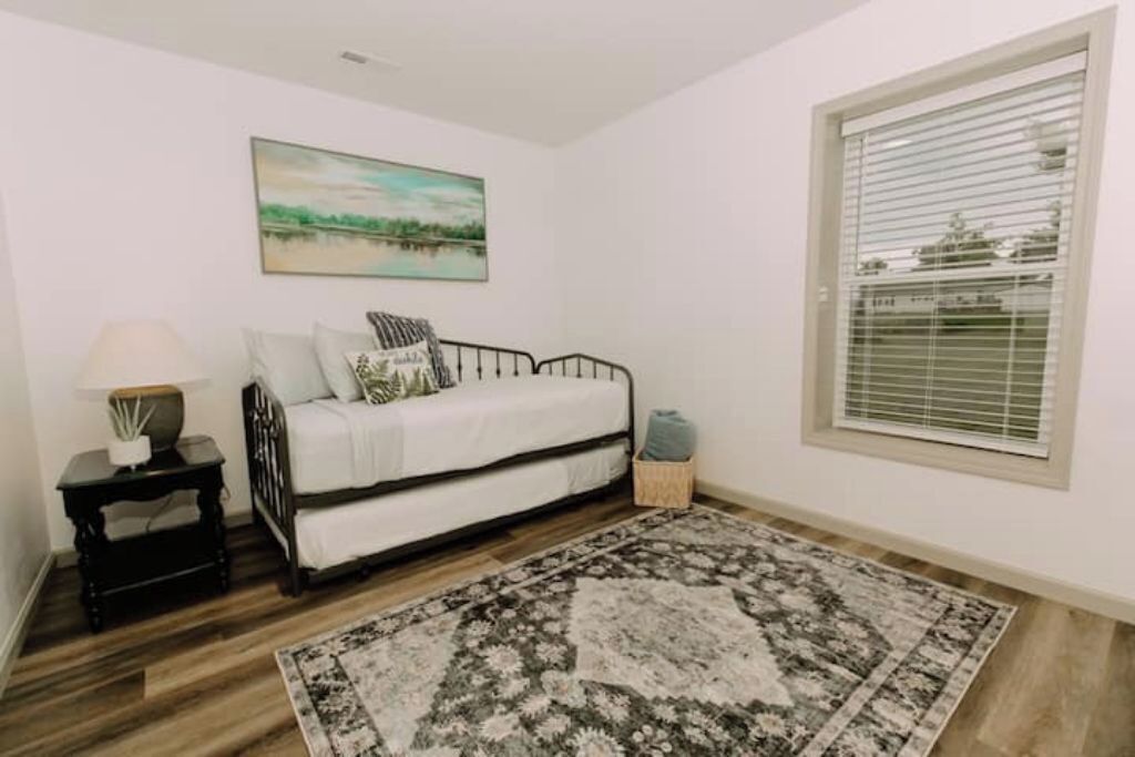 Une pièce avec un canapé blanc, un tapis à motifs, une petite table avec une lampe et une fenêtre avec des stores.