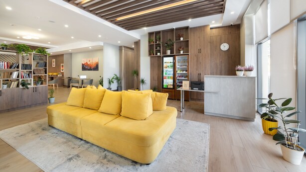 Una sala moderna con un sofá amarillo, techo de madera y una cocina con refrigerador y alacenas.