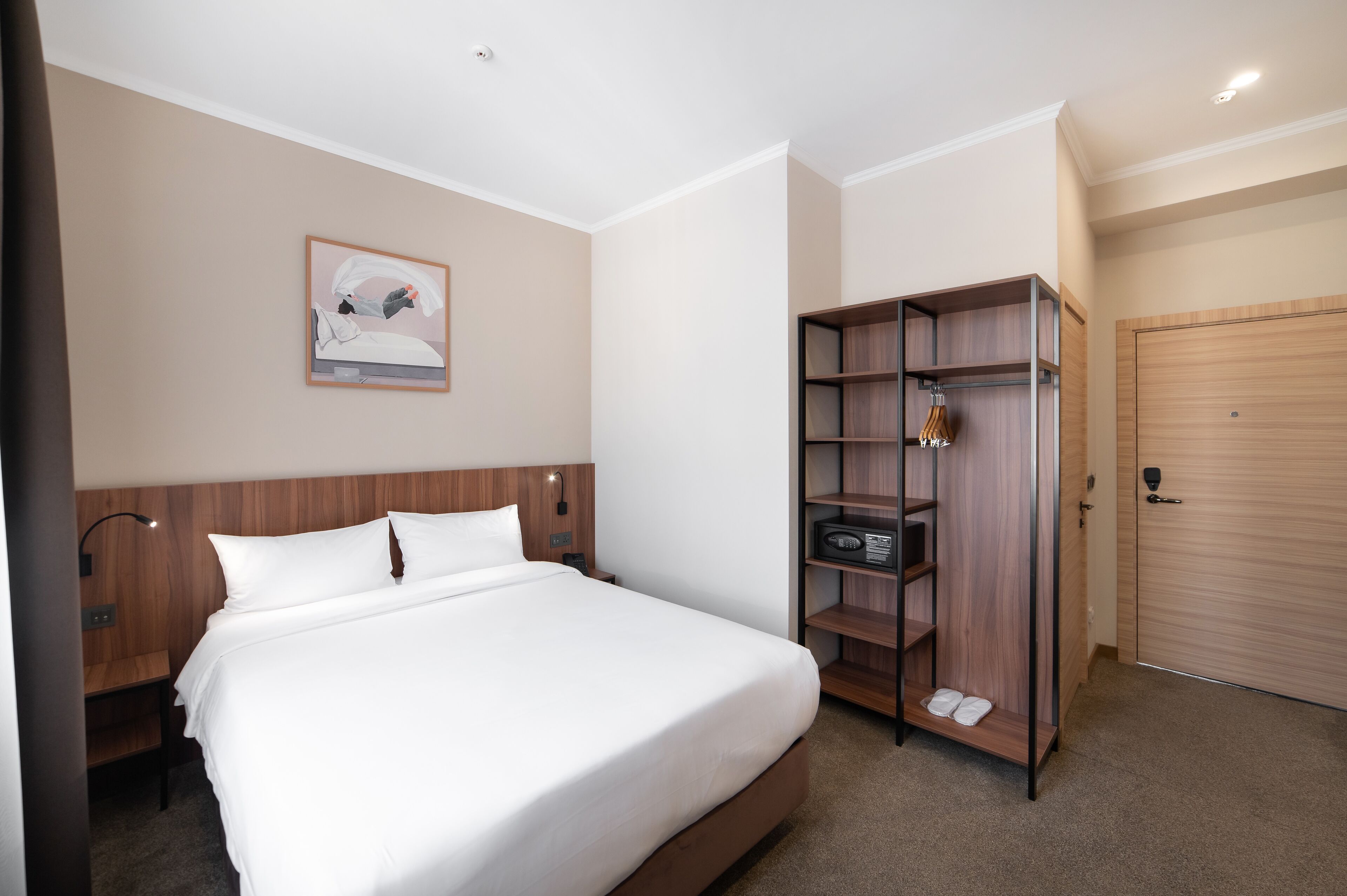 A hotel room with a large bed, a wooden wardrobe, a bedside table, a lamp, and a framed picture on the wall.