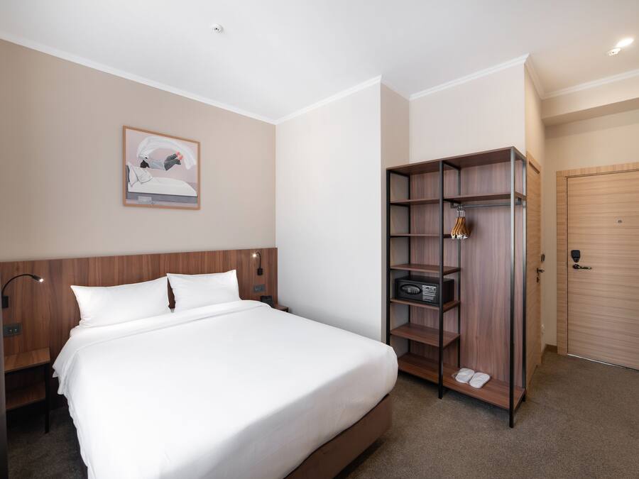 A hotel room with a large bed, a wooden wardrobe, a bedside table, a lamp, and a framed picture on the wall.