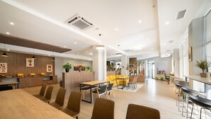 Una sala de conferencias moderna y bien iluminada con mesas y sillas de madera, una zona de cocina con sillas amarillas y un área de comedor con sofás amarillos.