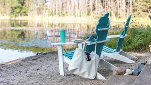 Dos sillas Adirondack con un esquema de colores blanco y aguamarina, un bolso blanco y una taza aguamarina sobre una playa arenosa con un lago rodeado de bosque al fondo.