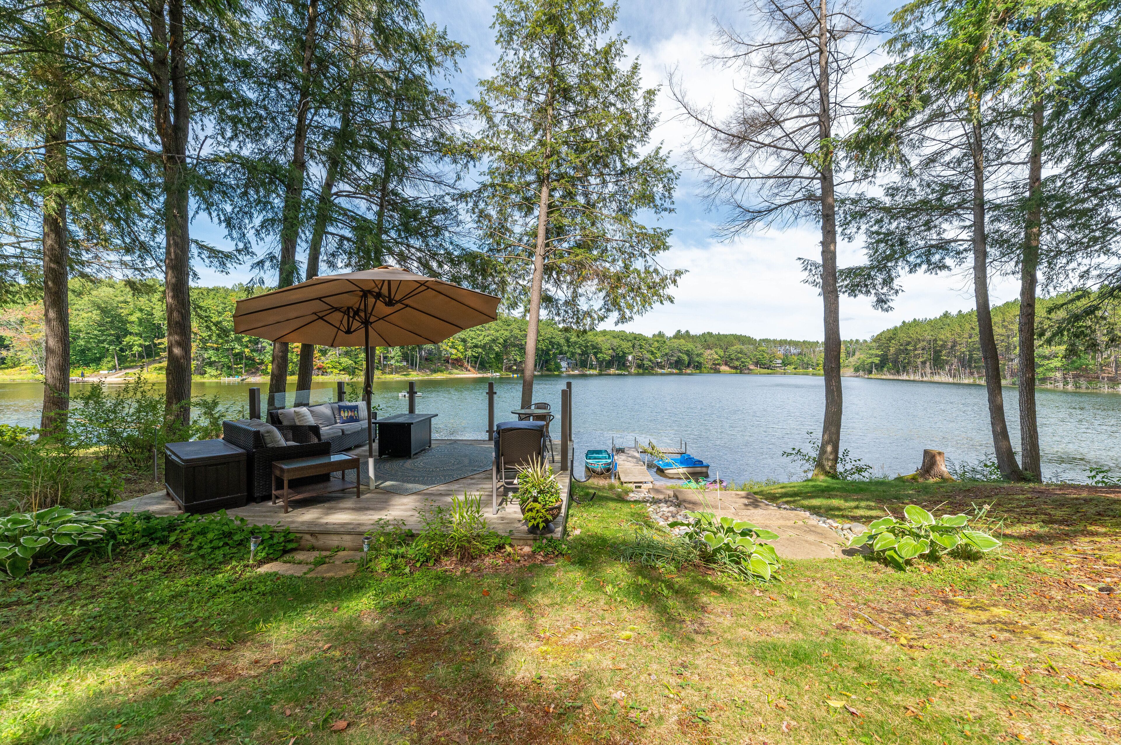 A lakeside patio with outdoor furniture and a large umbrella, surrounded by tall trees and a calm lake.
