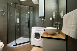 A modern bathroom with a glass-enclosed shower, a built-in washing machine, and a large sink with a mirror above. - Comfy Apt. w/ View – Lagos (Lagos)