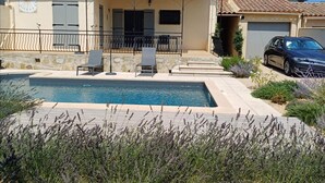 A house with a swimming pool, two lounge chairs, and a car parked in the driveway.