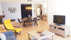 A living room with a yellow chair, a blue sofa, a TV on a stand, a dining area with a table and chairs, and a wall with a motivational quote.