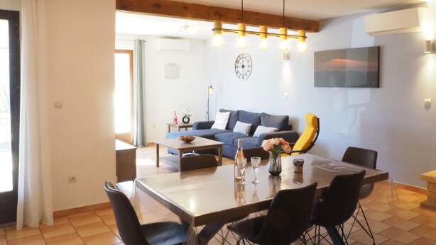 A modern living room with a sofa, dining table, and chairs. There is a clock on the wall and a painting above the sofa.