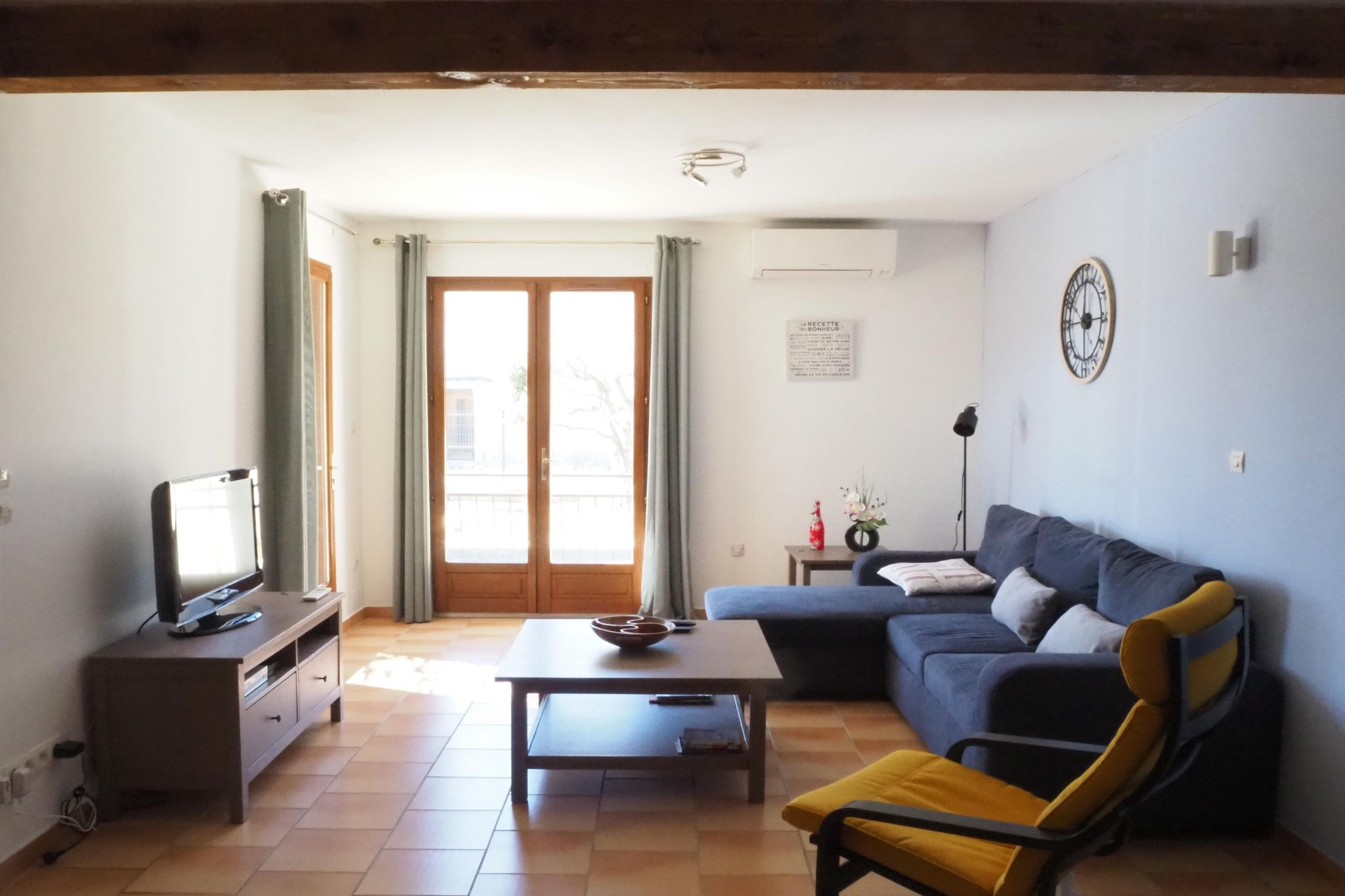 A living room with a television, sofa, coffee table, and a chair. There is a window with curtains, a wall clock, and a small table with a vase.