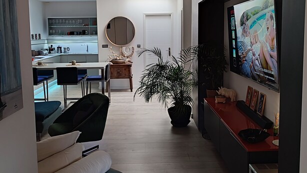 Una sala de estar moderna con sofá, televisor montado en la pared, comedor con sillas y mesa, y cocina al fondo.