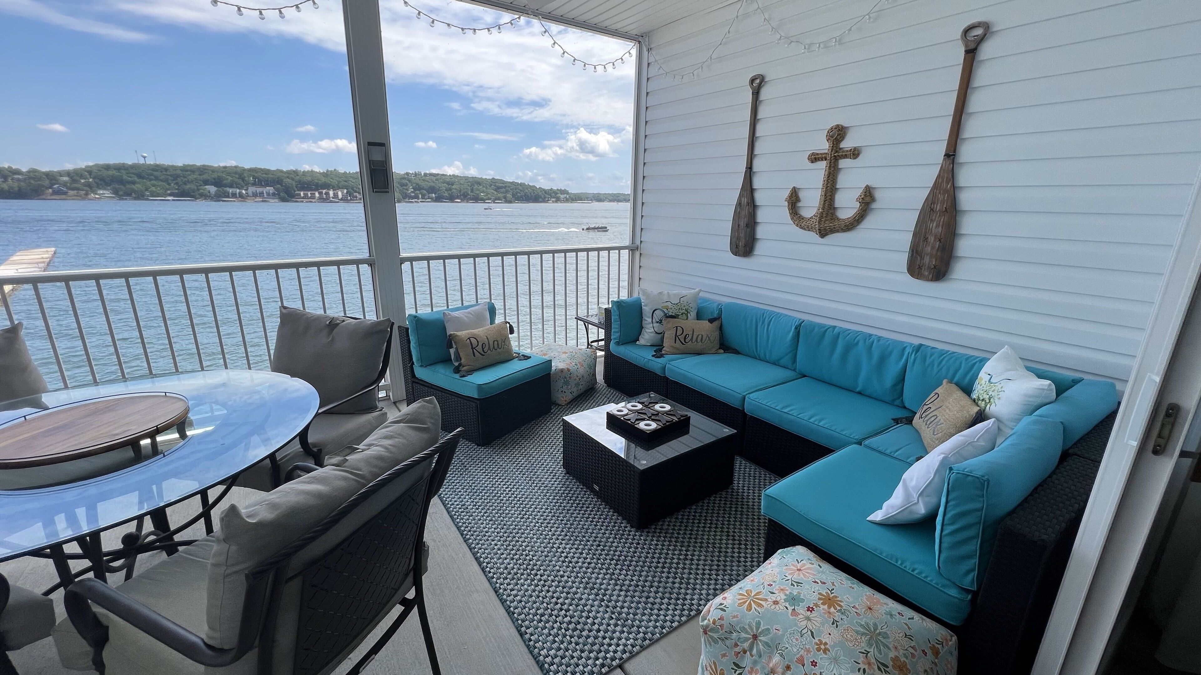 A covered outdoor seating area with a view of the water, a sofa, chairs, a table, and decorative anchors on the wall.