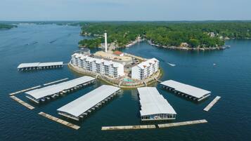 A waterfront hotel with multiple docked boats, a central building with a swimming pool, and a forested island in the background.