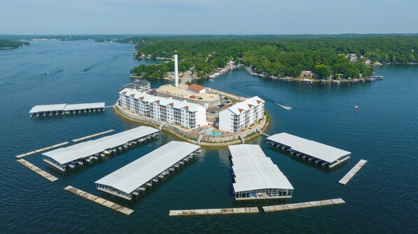 A waterfront hotel with multiple docked boats, a central building with a swimming pool, and a forested island in the background.