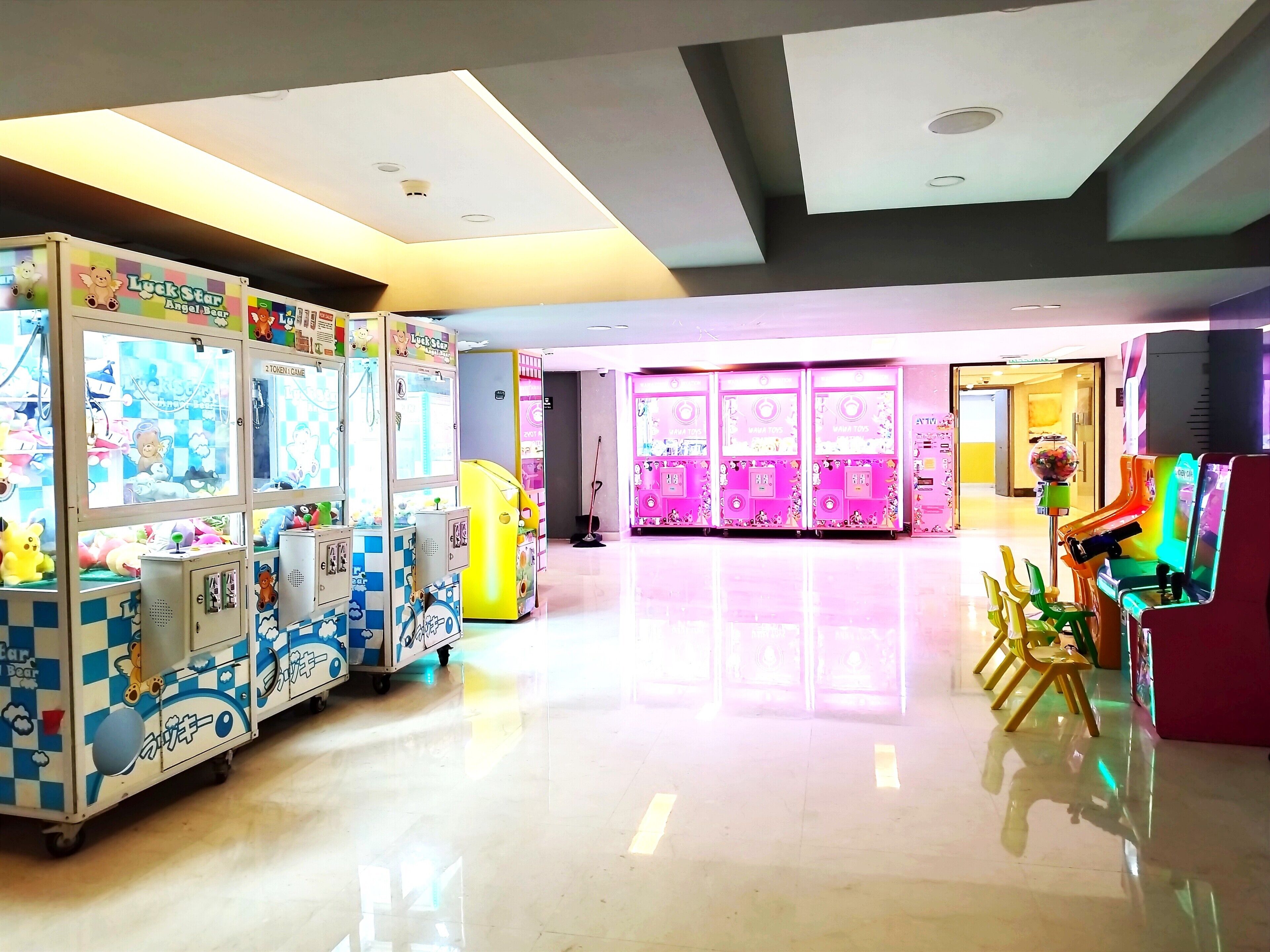A brightly lit indoor area with arcade games, including claw machines and racing games, and a few chairs near the entrance.