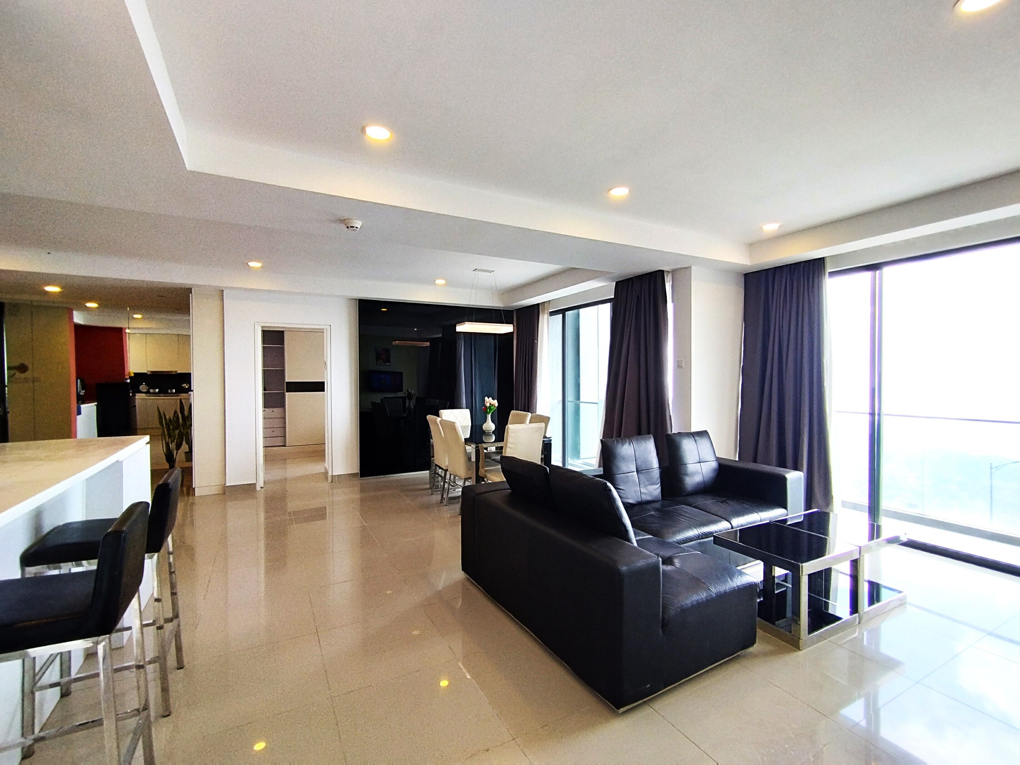 A modern living room with a black leather sofa, a dining area with chairs, a kitchen in the background, and large windows.