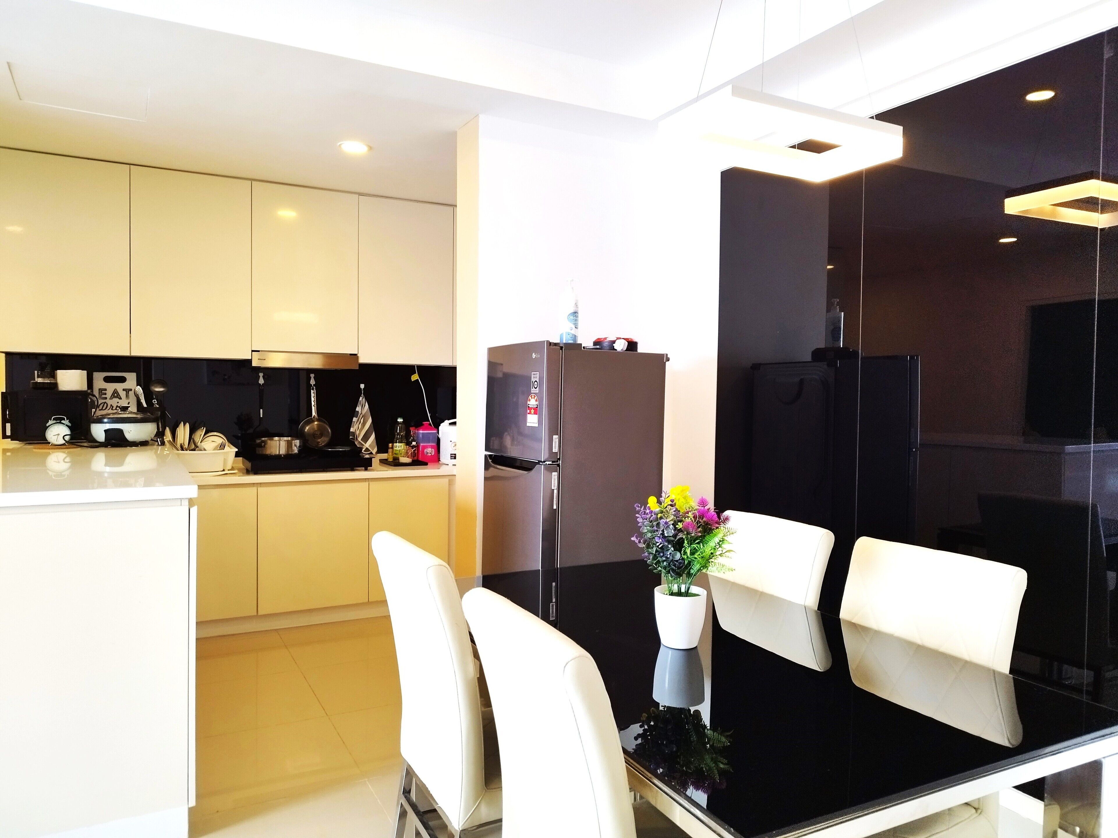 A modern kitchen with white cabinets, a black countertop, and a stainless steel refrigerator. There is a dining area with white chairs and a glass table.