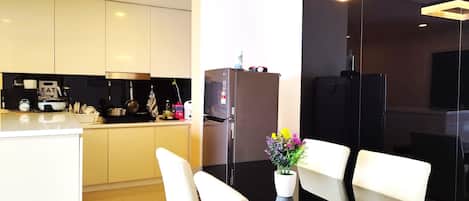 A modern kitchen with white cabinets, a black countertop, and a stainless steel refrigerator. There is a dining area with white chairs and a glass table.