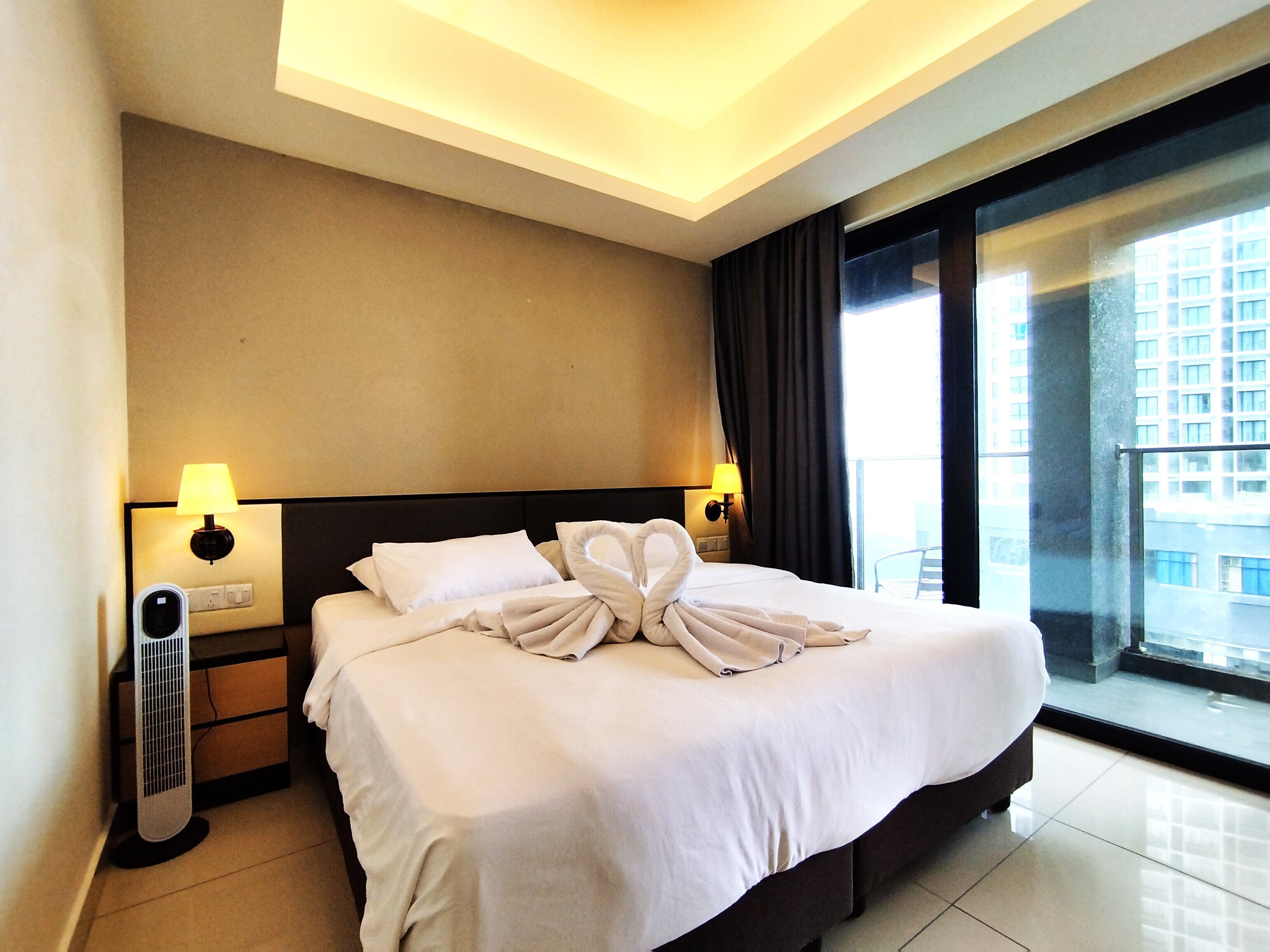 A hotel room with a large bed, two bedside lamps, a heater, and a view of a building through a large window.