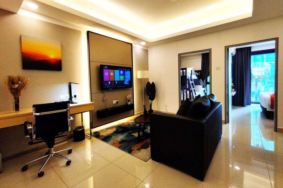 A modern hotel room with a flat-screen TV mounted on the wall, a black sofa, a desk with a computer, and a small plant.
