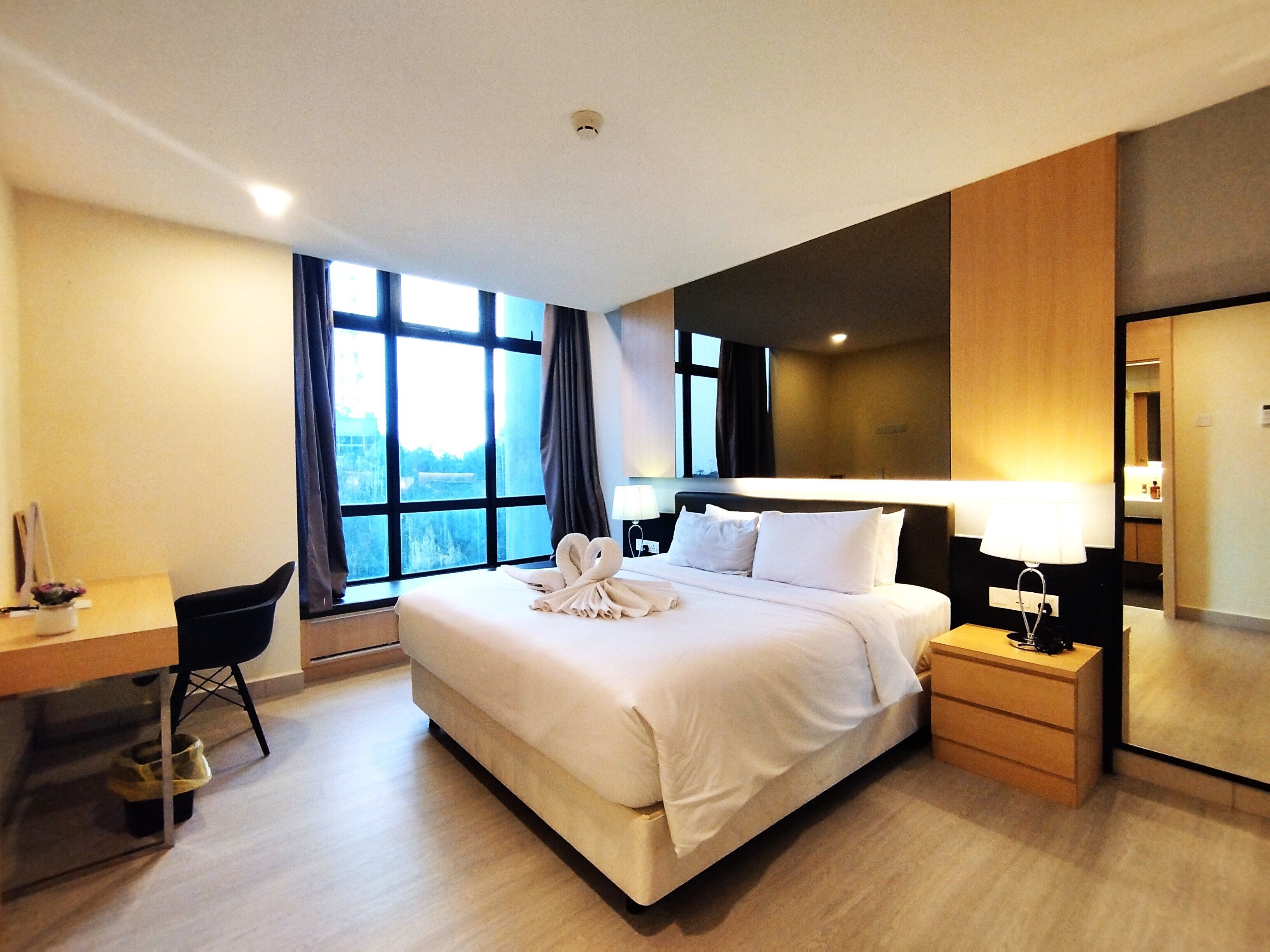 Quarto de hotel com cama, mesa de cabeceira, escrivaninha, cadeira e uma janela grande.