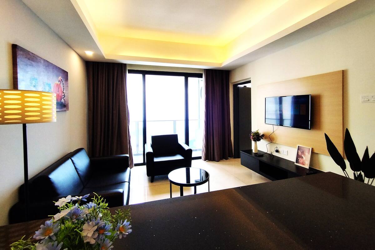 A modern hotel room with a sofa, a dining table, a TV, and a balcony with curtains.