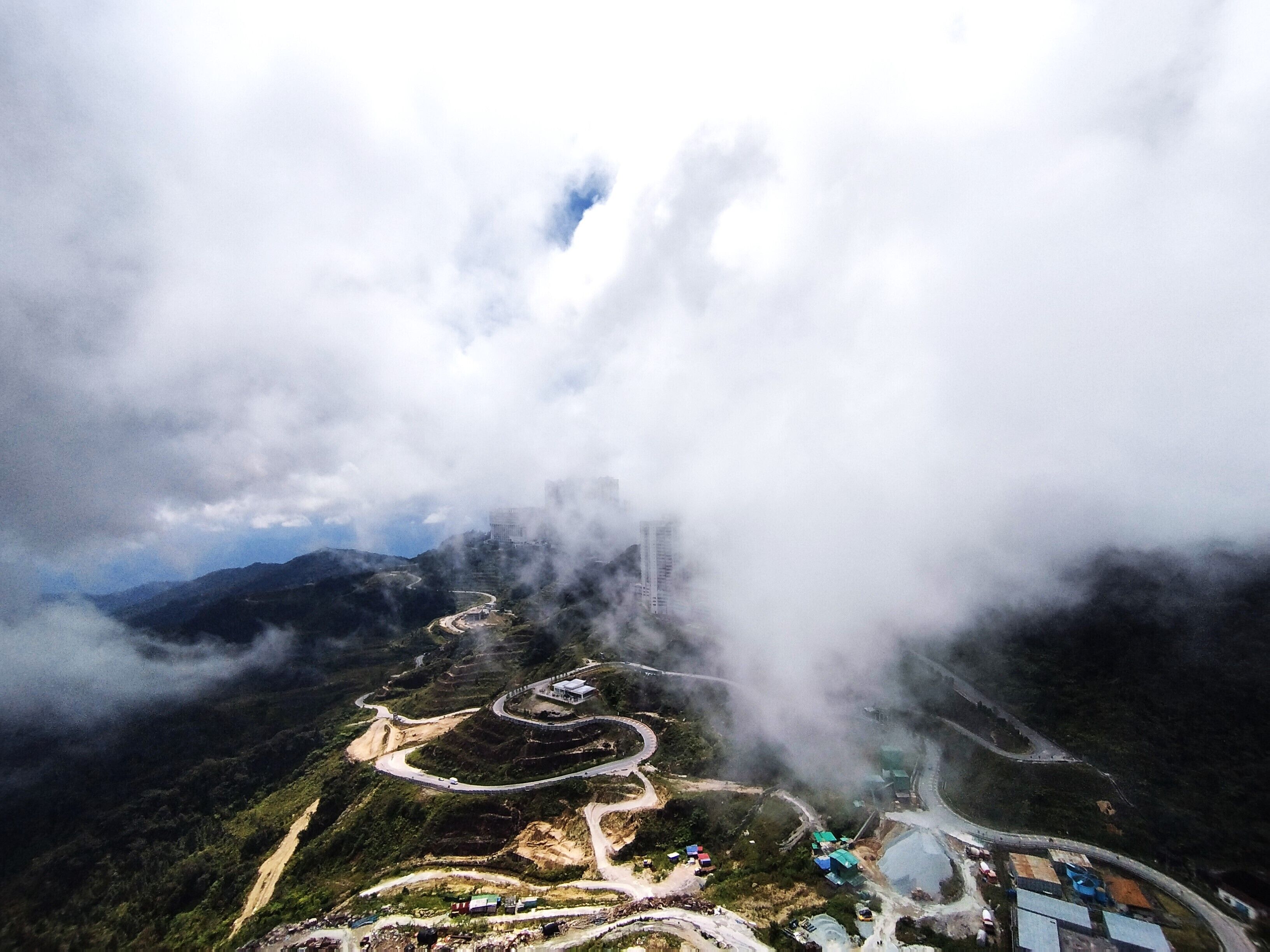An aerial view of a mountainous area with winding roads, buildings, and a large cloud-covered mountain peak.