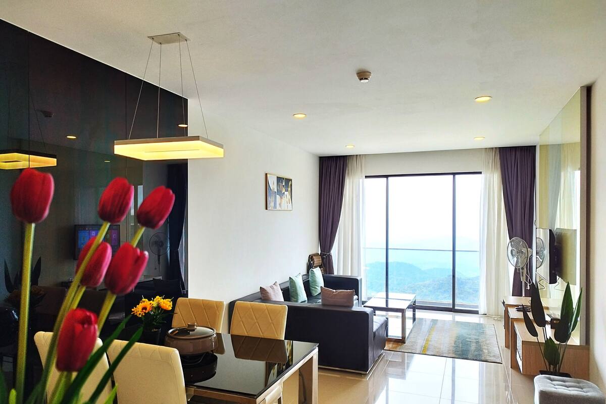 A modern living room with a sofa, dining table, and chairs. There is a large window with curtains, a ceiling fan, and a view of the mountains.